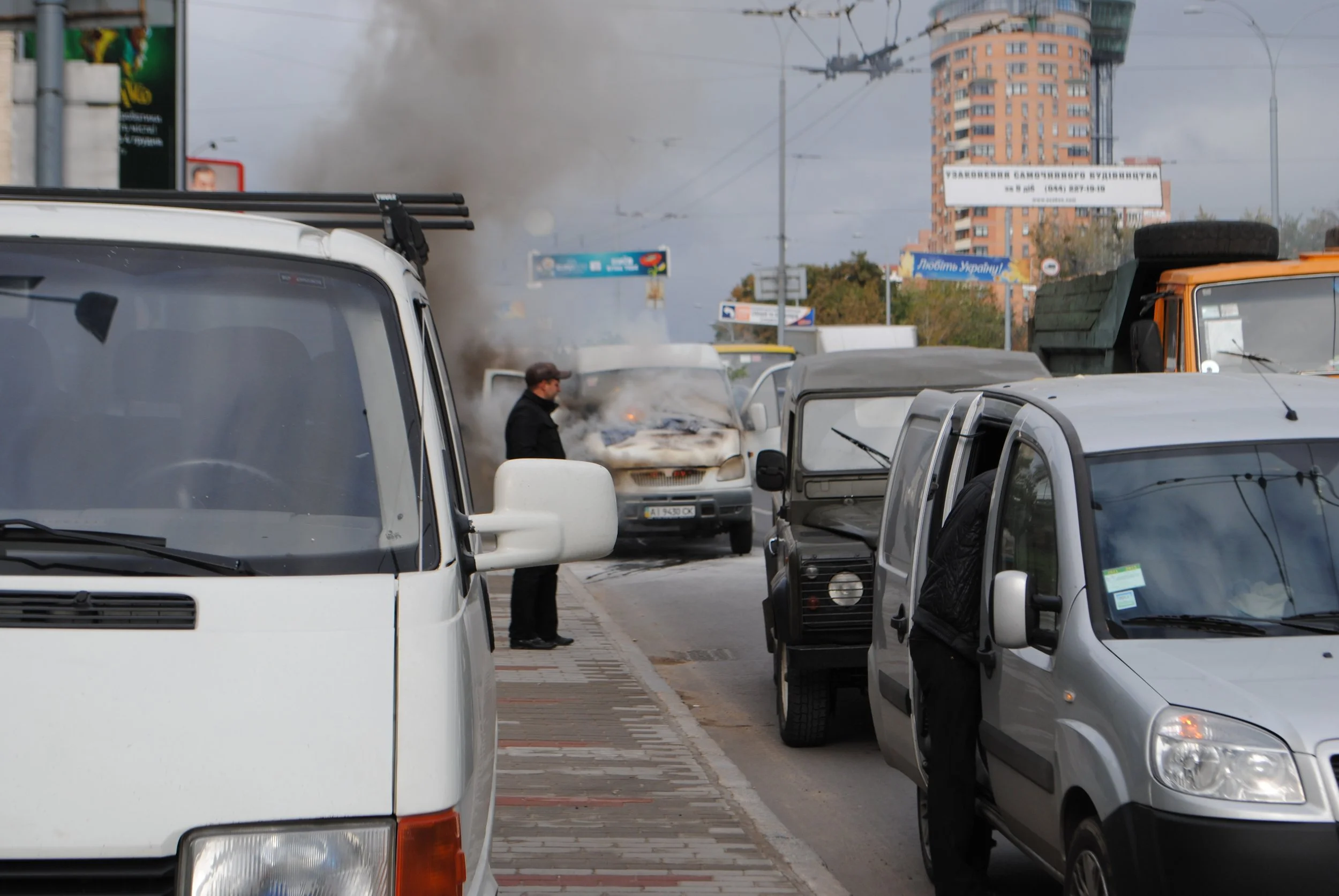 A busy city street with several vehicles including vans and cars, with one vehicle emitting thick black smoke. A person stands near the vehicles, and high-rise buildings and billboards are visible in the background.