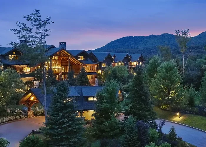 A mountain resort with multiple wooden cabins illuminated at dusk, surrounded by lush green trees and a winding driveway.