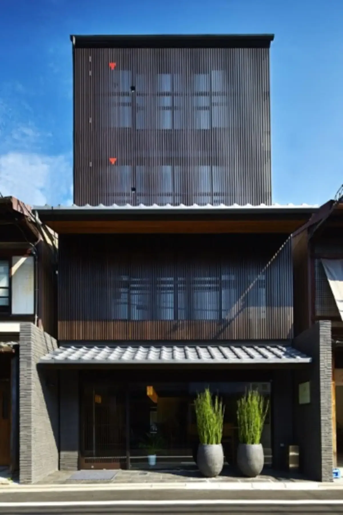 Modern multi-story house with black exterior, large windows, and two potted plants at the entrance, under a blue sky.