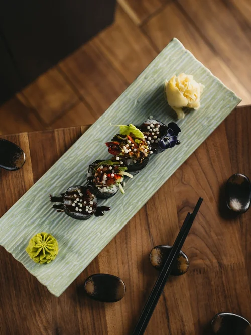 Sushi rolls garnished with sauce, scallions, and sesame seeds, accompanied by wasabi and soy sauce in small bowls on a long plate, on a wooden table.