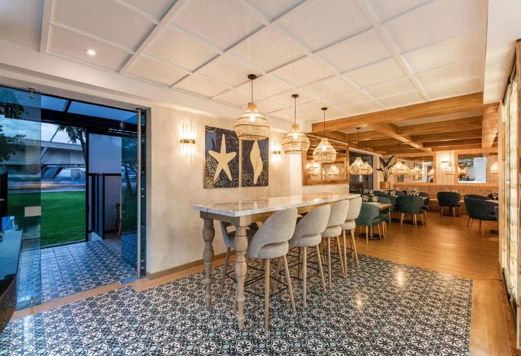 Interior of a modern restaurant with a bar area, hanging wicker lights, wooden ceiling, patterned tile floor, and seating with green and gray chairs, large windows, and outdoor view.