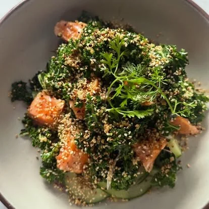 A bowl of cooked salmon and broccoli topped with sesame seeds and garnished with fresh herbs.