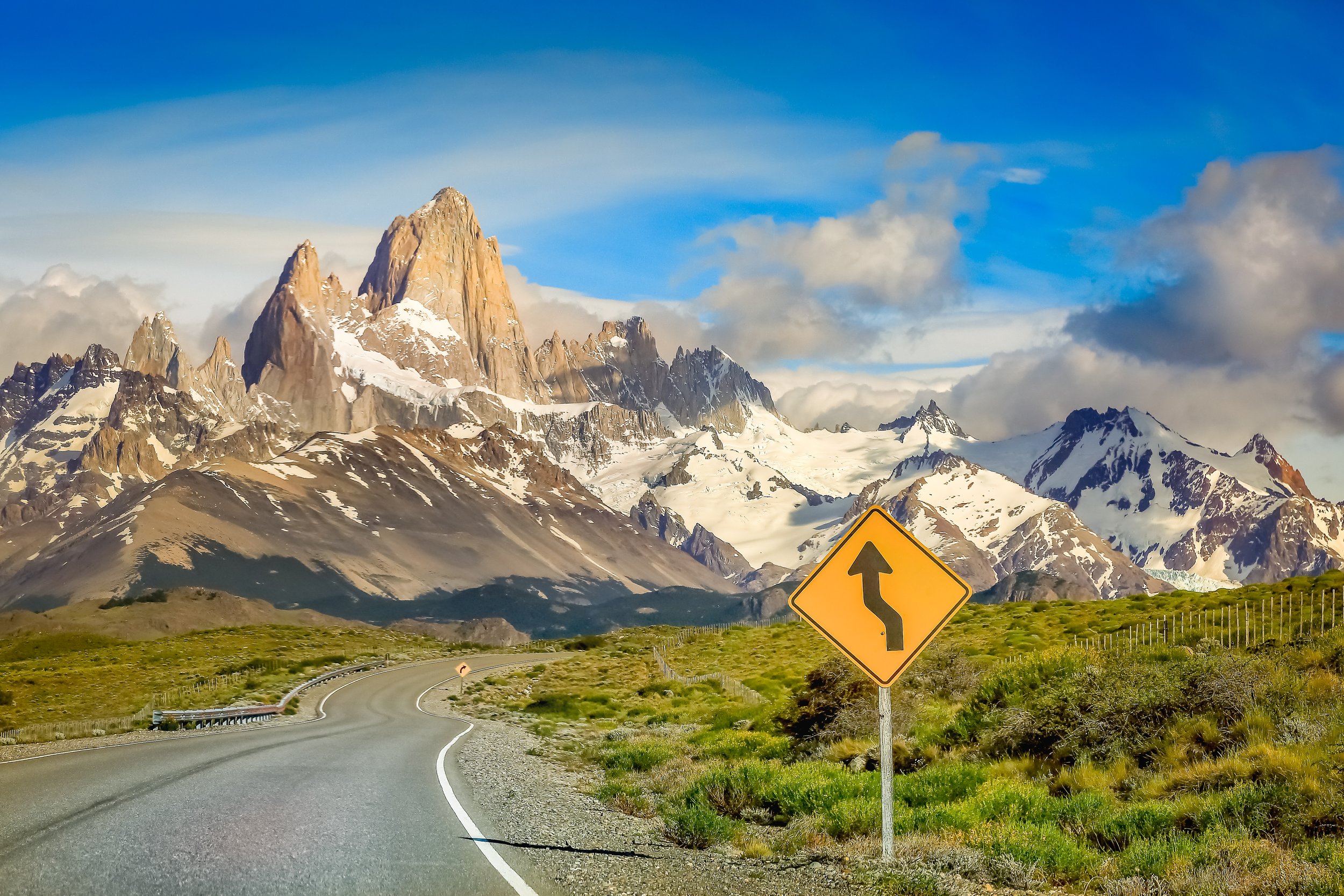 Scenic mountain road with a yellow sign indicating a left curve, snow-capped mountains in the background, and a partly cloudy sky.