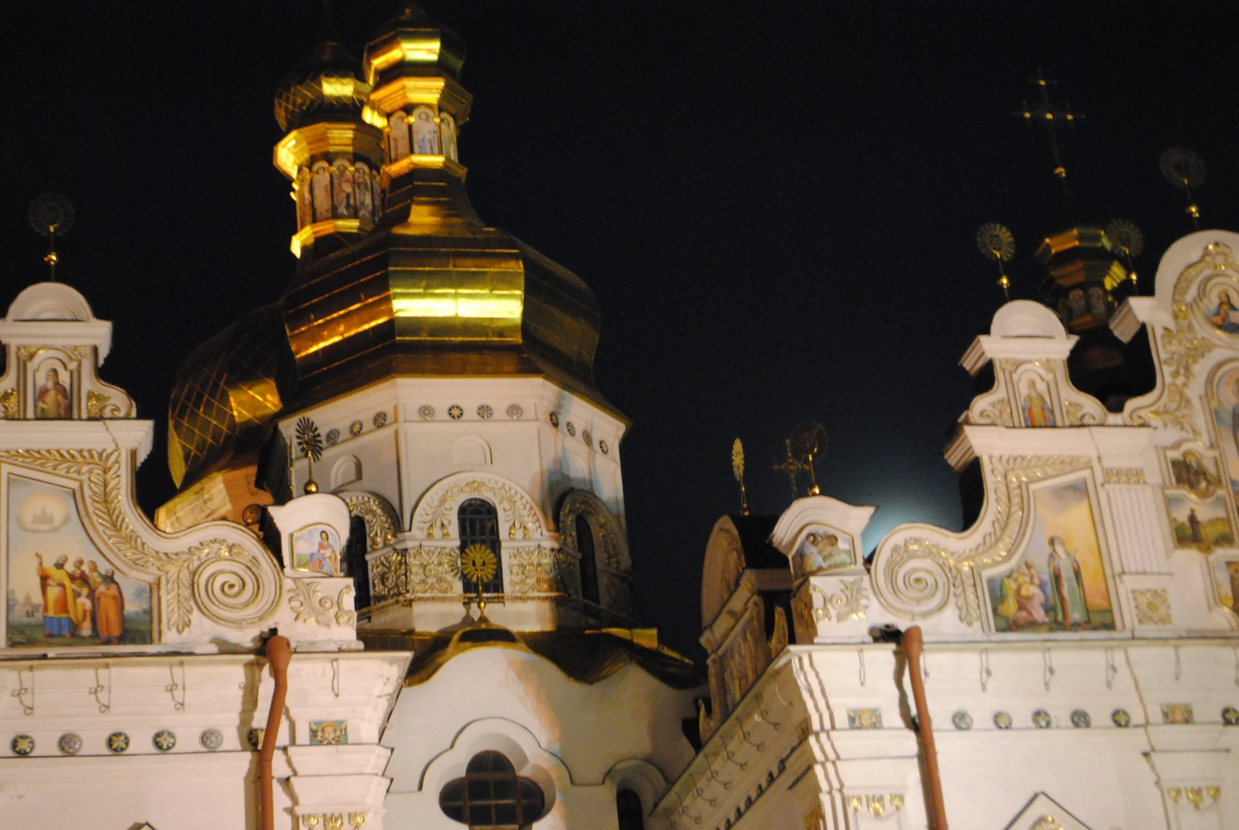 Nighttime view of a decorated church with golden domes, white walls, and religious icons, illuminated against a dark sky.