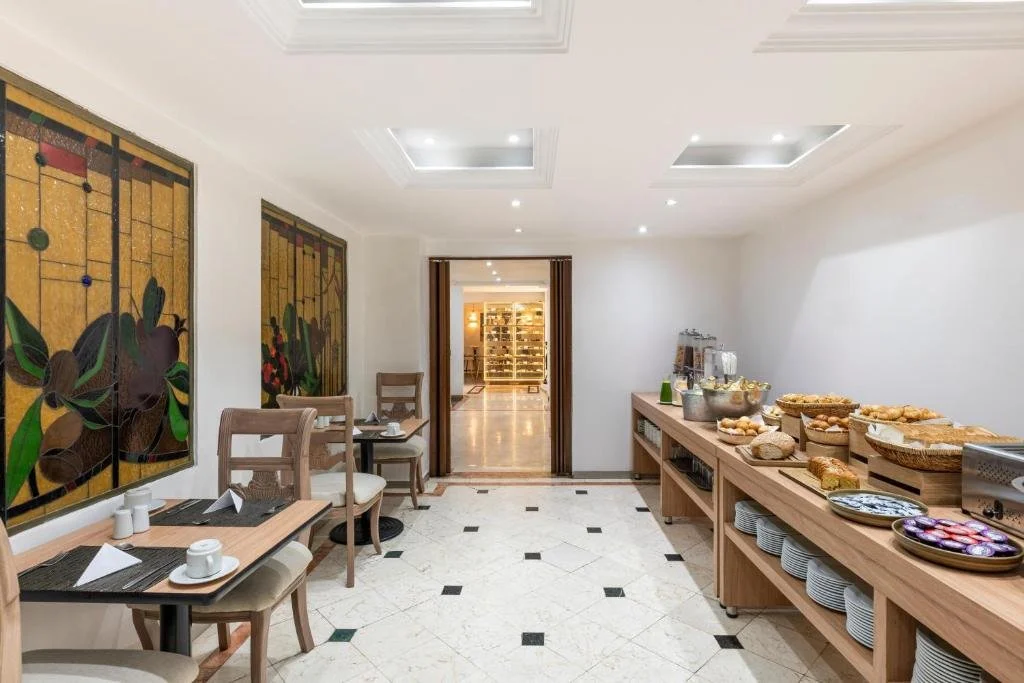 A breakfast buffet setup in a dining room with wooden tables, chairs, and a side table with food items including bread, pastries, and plates, with decorative wall art and a doorway leading to another room.