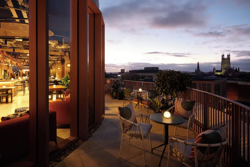 Cityscape viewed from a rooftop bar at sunset, with outdoor seating and potted plants.