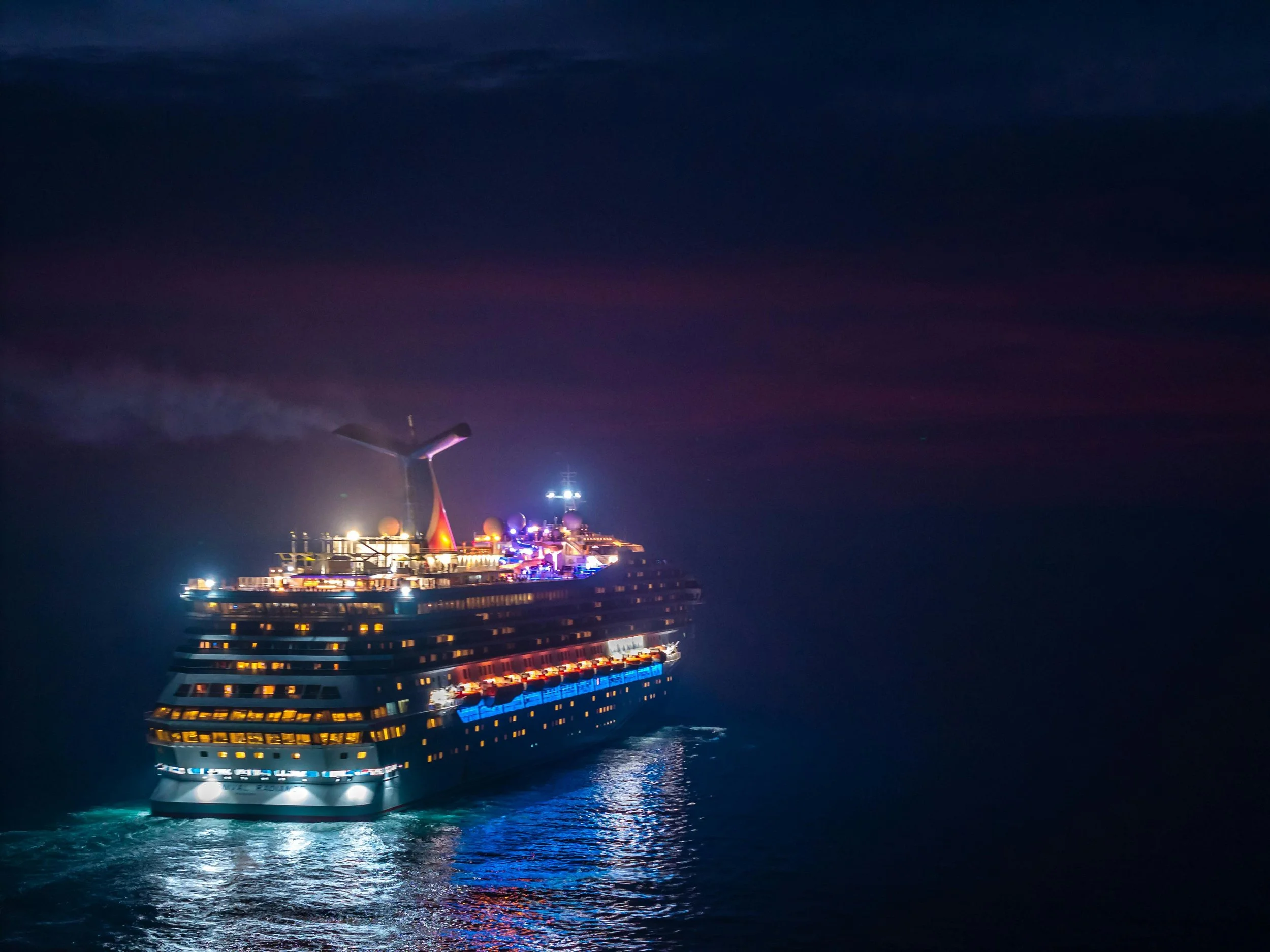 A large cruise ship docked at night with colorful fireworks lighting up the sky above.