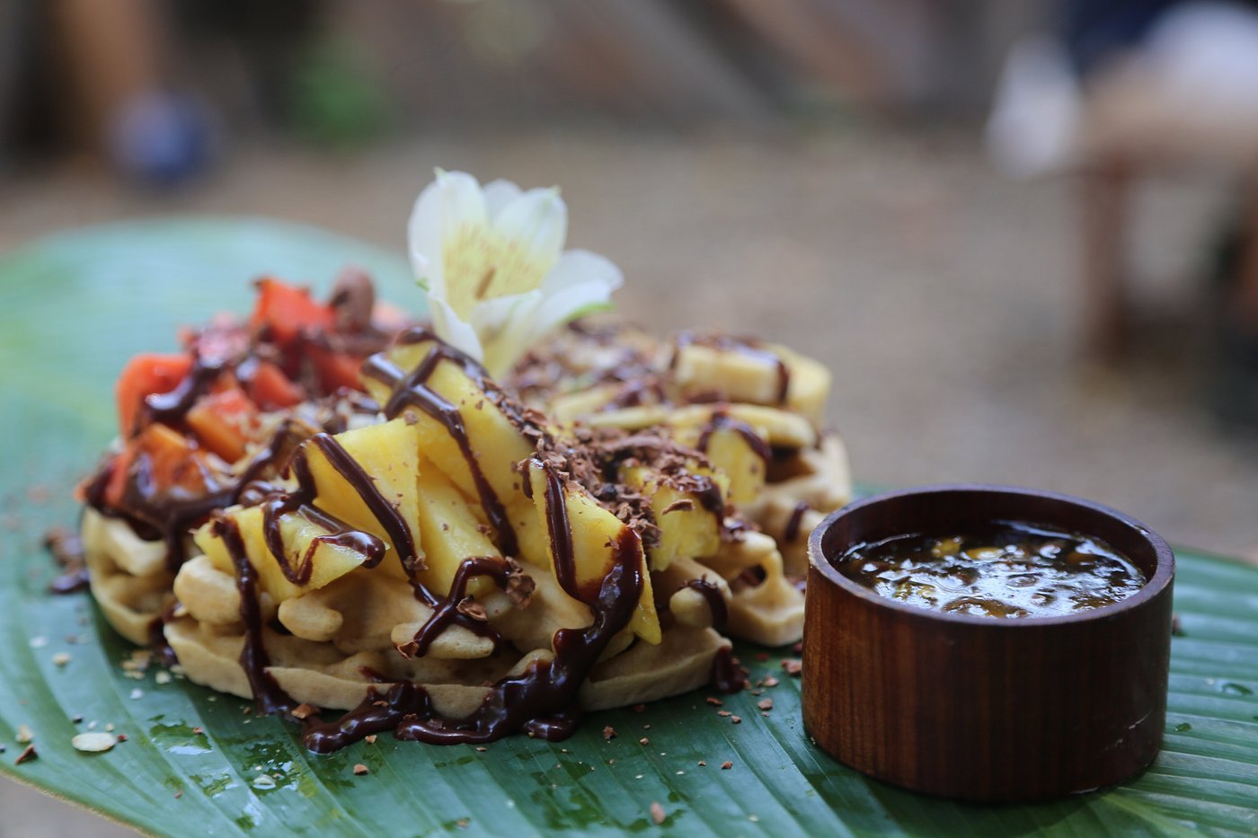 Waffle topped with sliced bananas, chocolate drizzle, and a flower, served with a small wooden bowl of syrup on a large green leaf.