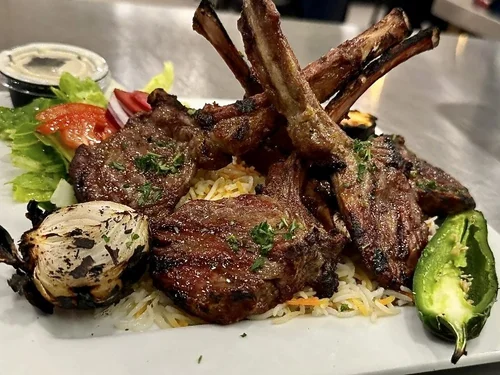 A plate of grilled lamb chops served over rice with a side of grilled green chili, roasted garlic, fresh lettuce, tomato slices, and salad dressing.