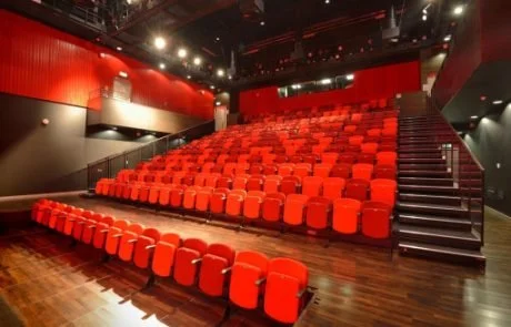 Theater auditorium with red seats and black stairs, empty with dim lighting.