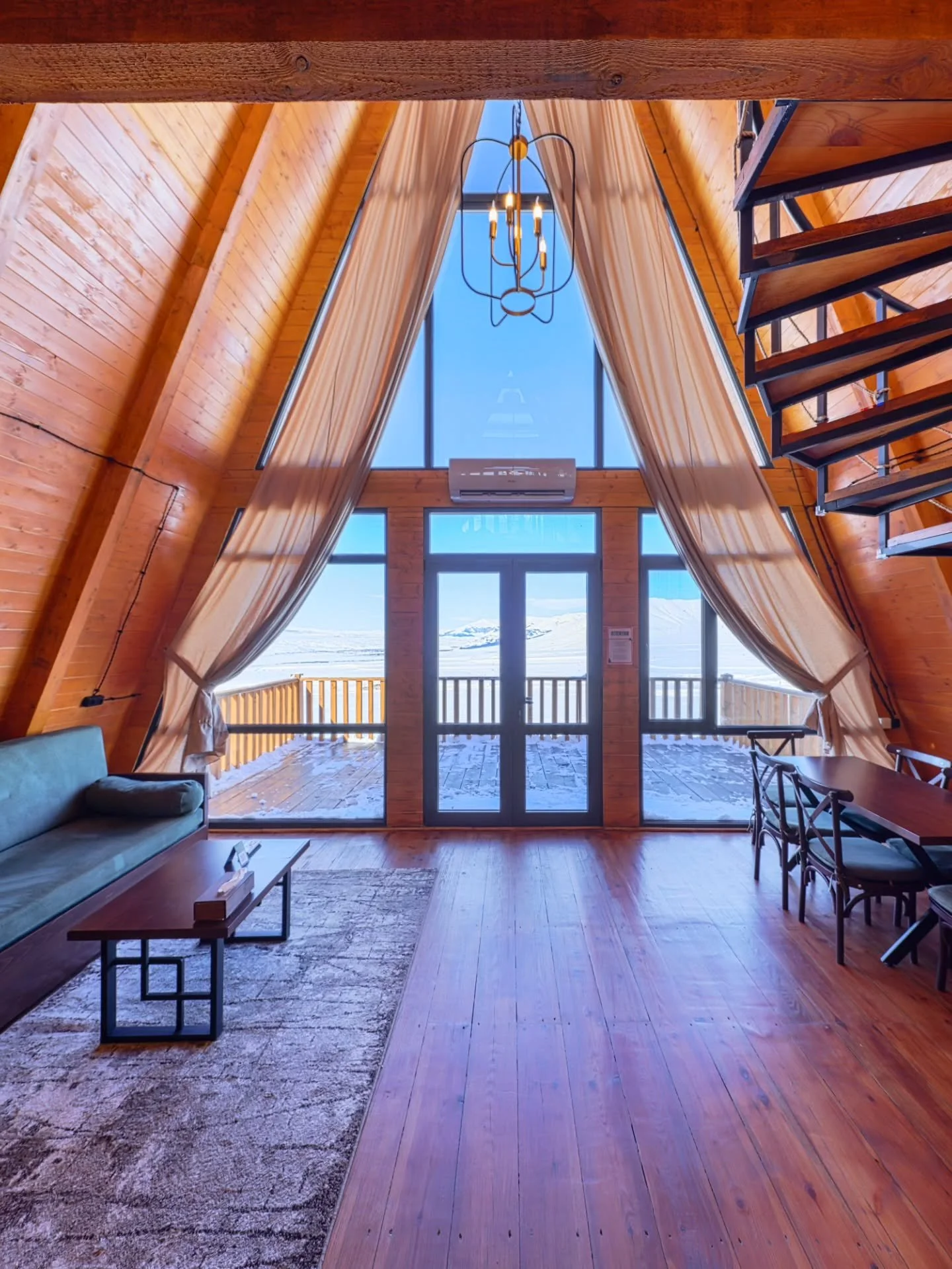 A cozy A-frame cabin living room with wooden walls and floor, large window with curtains, a chandelier, a couch, a coffee table, a dining table with chairs, and a loft staircase overlooking a snowy landscape outside.