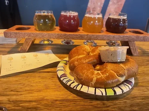 A plate with a bagel and a container of cream cheese, four small glasses of different flavored craft beer on a wooden table, and a tasting notes sheet.