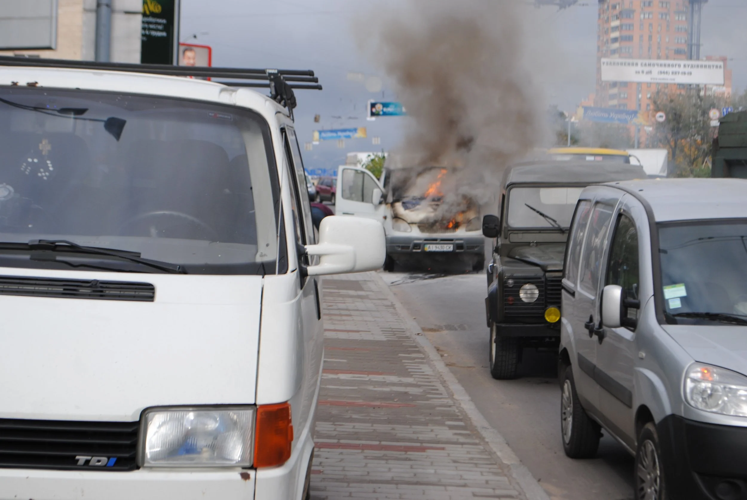 A white van on the street is on fire with black smoke coming from the engine area. Other vehicles are parked nearby, and urban buildings and billboards are visible in the background. Kiev November 2011, Marina Parrish ©