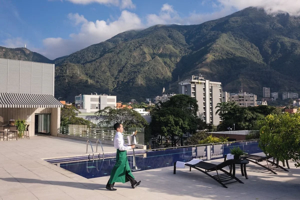 A person in a white shirt and green apron walking beside a swimming pool on a rooftop with city buildings and green mountains in the background.