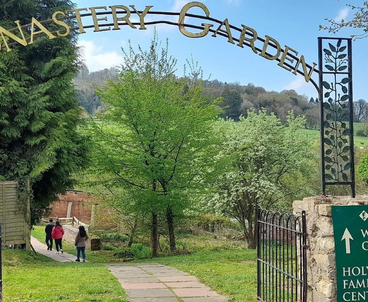 Entrance to Nastery Garden with a metal archway, lush green trees and a stone pathway, three people walking in the garden, rolling green hills in the background, cloudy sky.