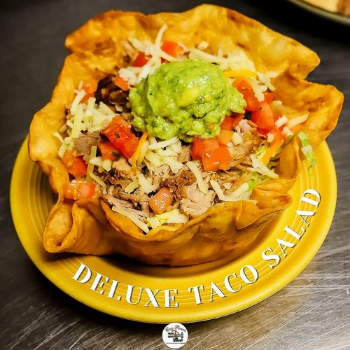 Taco salad served in a crispy tortilla bowl topped with guacamole, shredded cheese, diced tomatoes, shredded lettuce, and ground meat, on a yellow plate with "DELUXE TACO SALAD" written on the rim.
