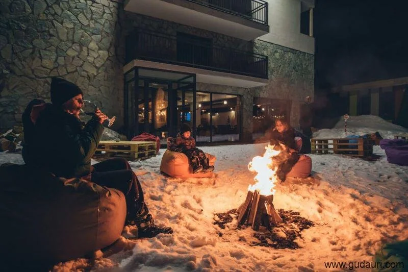 Four people sitting on bean bags around a campfire on snow outside a house at night.