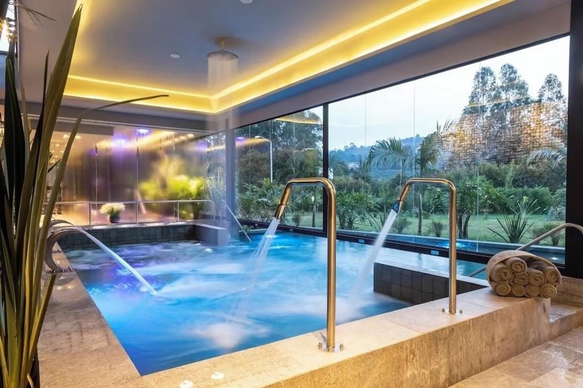 Indoor spa pool with jets, surrounded by large glass windows showing greenery outside, with rolled towels on the edge and soft yellow lighting on the ceiling.