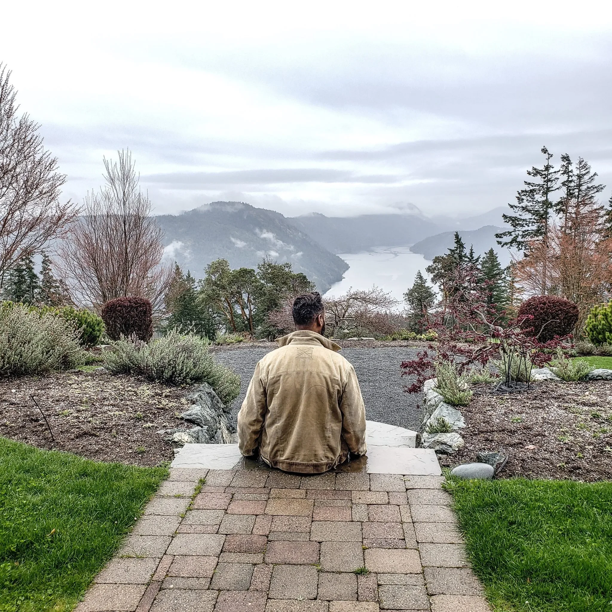 A man sitting on a brick pathway in a garden overlooking a lake and mountain landscape with cloudy skies.
