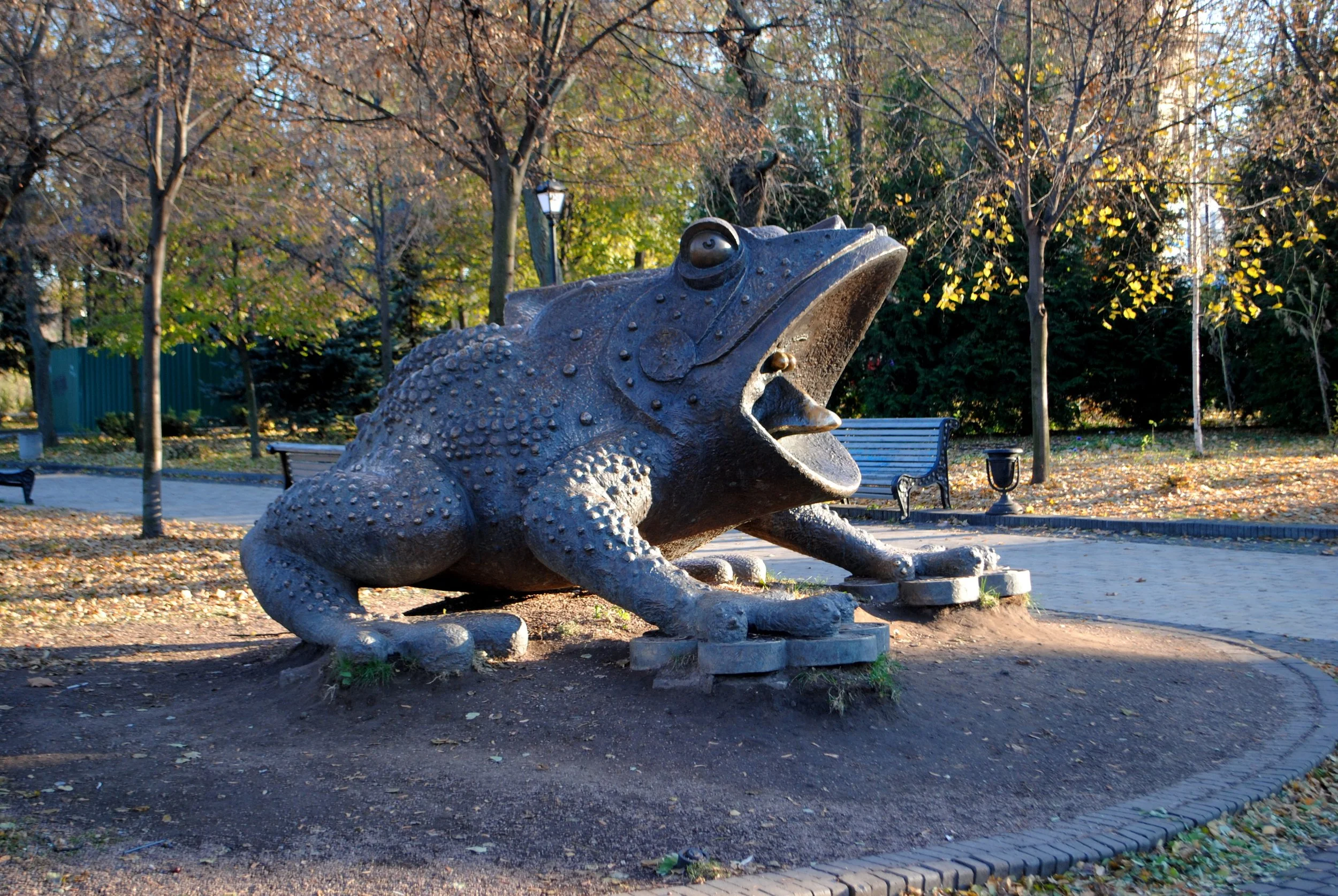 Sculpture of a large frog in a park with trees and fallen leaves.