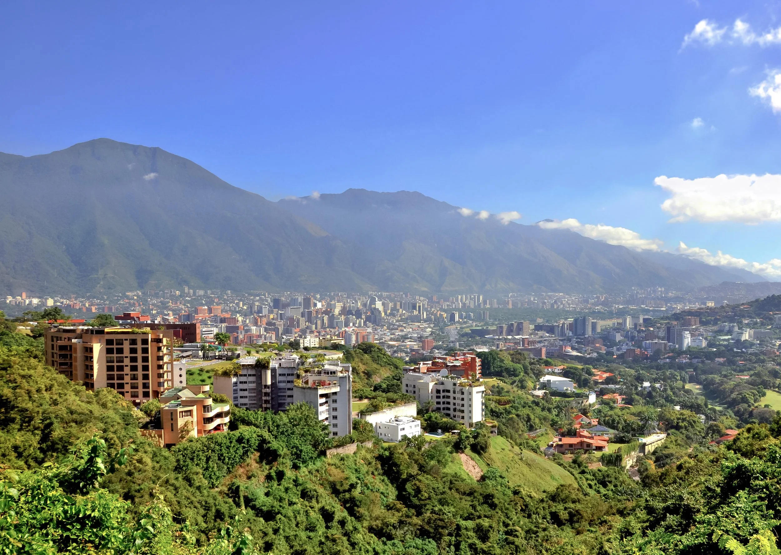 A cityscape with numerous buildings in the foreground, green hills on the sides, and large mountains in the background under a bright blue sky with some clouds.