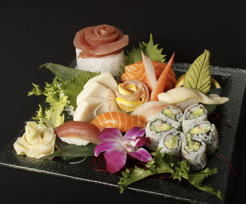 Assorted sushi platter with slices of raw fish, sushi rolls, garnishes, and decorative leaves on a glass plate.