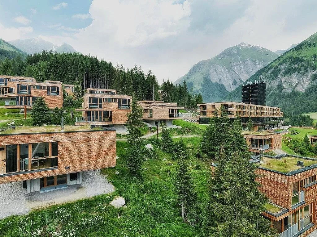 Modern wooden buildings on a hillside with green trees and mountains in the background, under a cloudy sky.