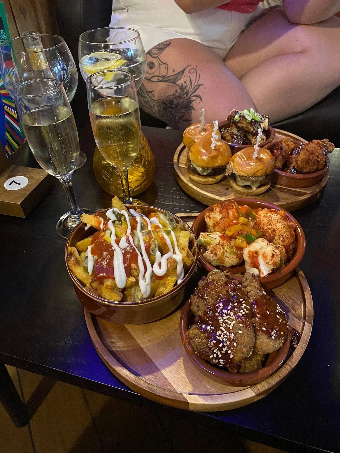 A table with various dishes including loaded fries topped with sauce and mayonnaise, crispy chicken wings with sesame seeds, sliders with cheese and toppings, and fried chicken pieces. There are four glasses of champagne and two bottles of what appea