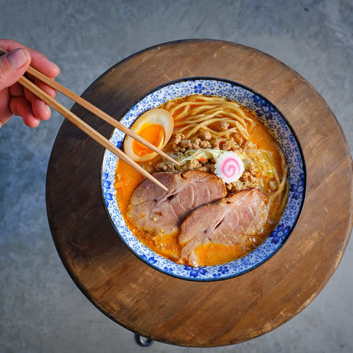 Bowl of ramen noodles with sliced pork, half boiled egg, narutomaki, minced meat, and vegetables on a wooden surface.