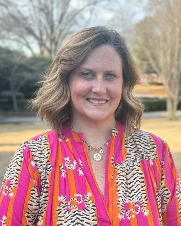 An elderly woman with short white hair, wearing a light green blouse and a beaded necklace, standing against a gray backdrop.