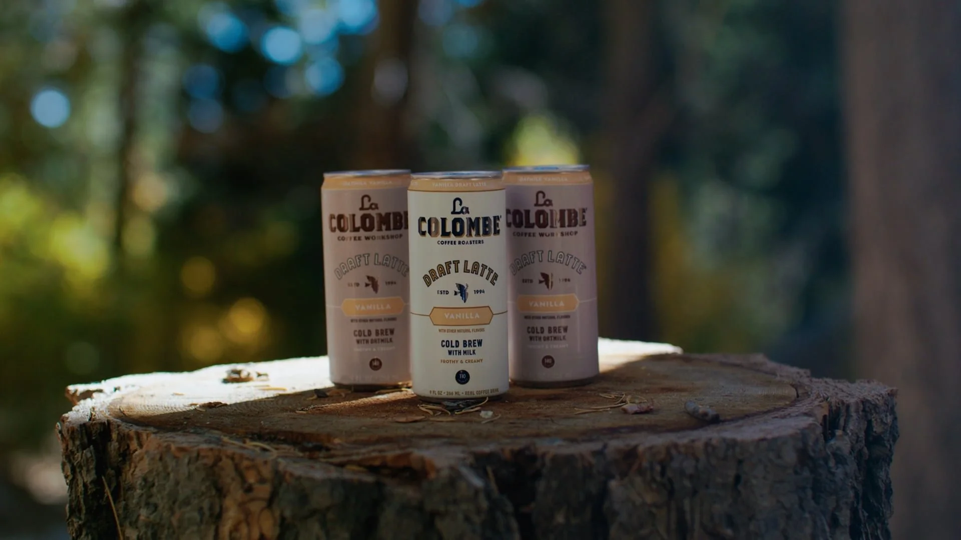 Three cans of La Colombe Draft Latte Vanilla flavor placed on a tree stump outdoors with blurred green forest background.