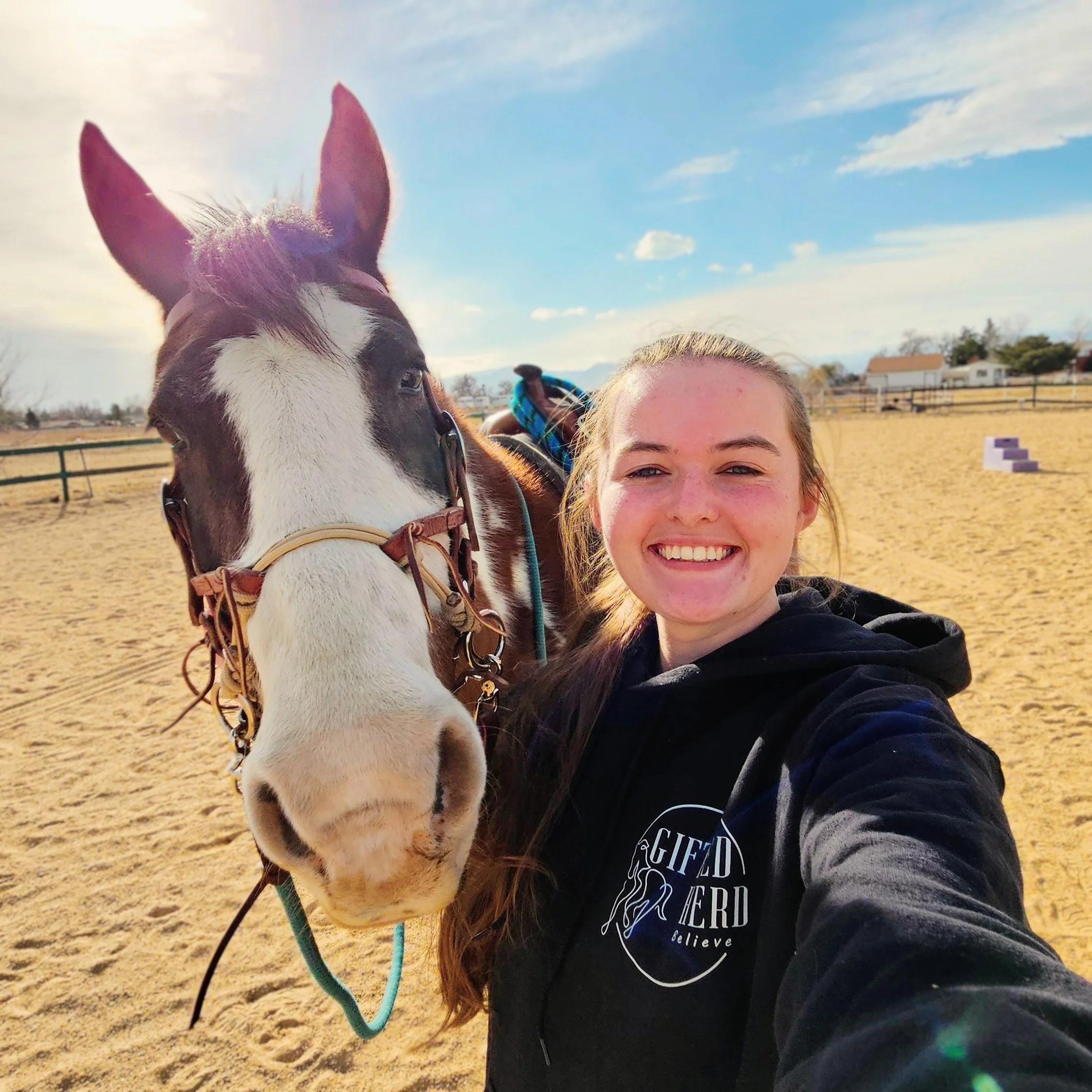 A young woman taking a selfie with a horse in an outdoor riding arena on a sunny day.