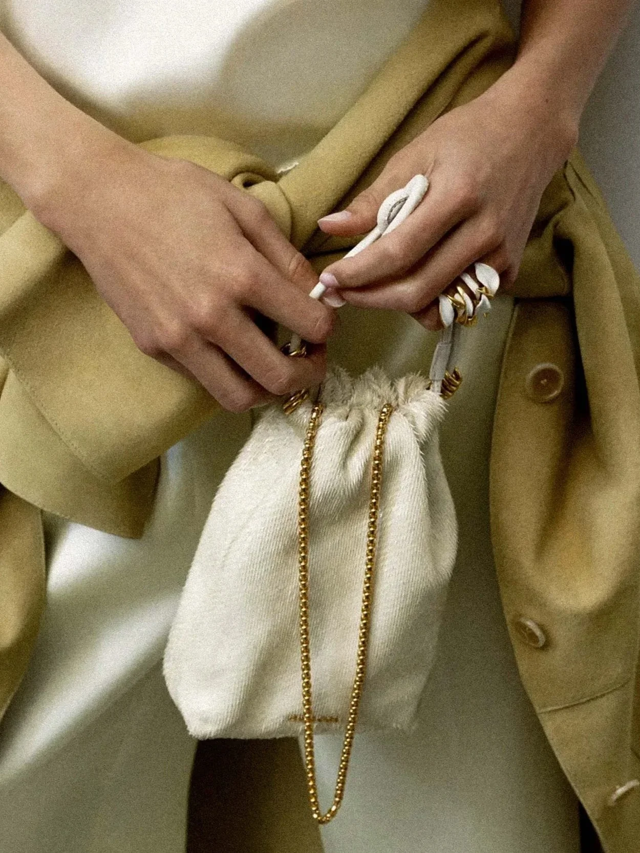 Close-up of a person’s hands adjusting the drawstring of a beige hoodie, with a beige coat over a white shirt and gold jewelry, including a chain necklace and rings.