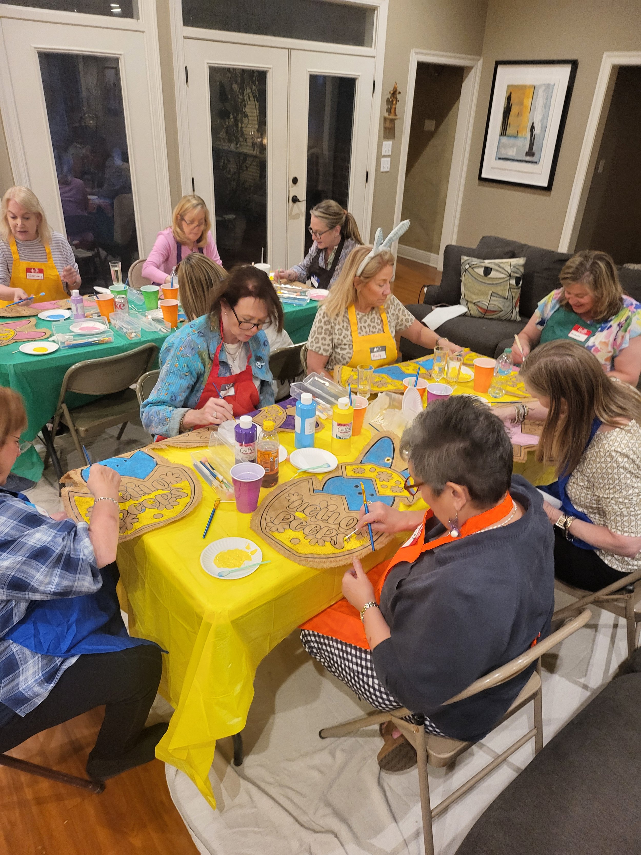 Group of women sitting around a table indoors, painting wooden plaques with colorful designs and the phrase 'Creative Crafters', at a craft party or workshop. Some women are wearing aprons and bunny ears.