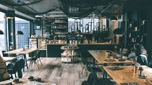Interior of a modern coffee shop with wooden tables, chairs, and shelves, and large windows letting in natural light.