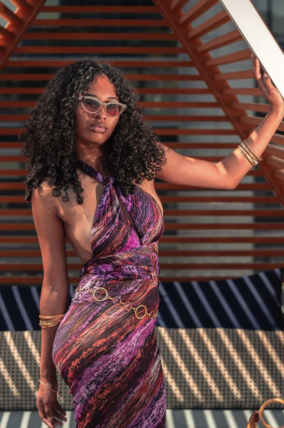 A woman with curly dark hair wearing stylish sunglasses and a colorful, patterned dress with a cutout side. She is accessorized with gold jewelry and standing outdoors against a modern, architectural background with orange and white slats.