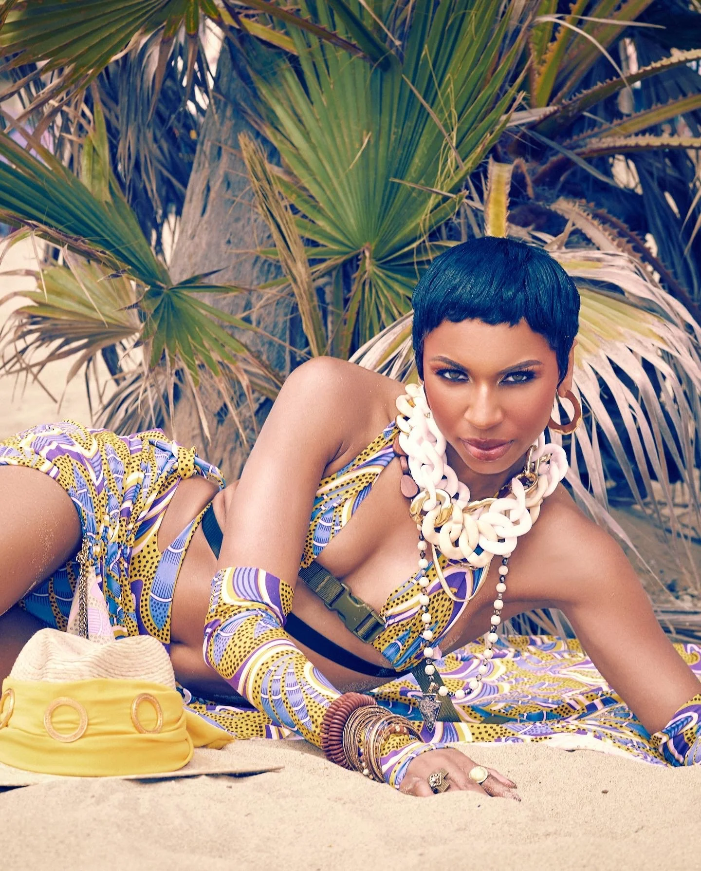 A woman with short dark hair lying on a sandy beach in front of tropical plants, wearing colorful patterned clothing, chunky jewelry, and a serious expression.