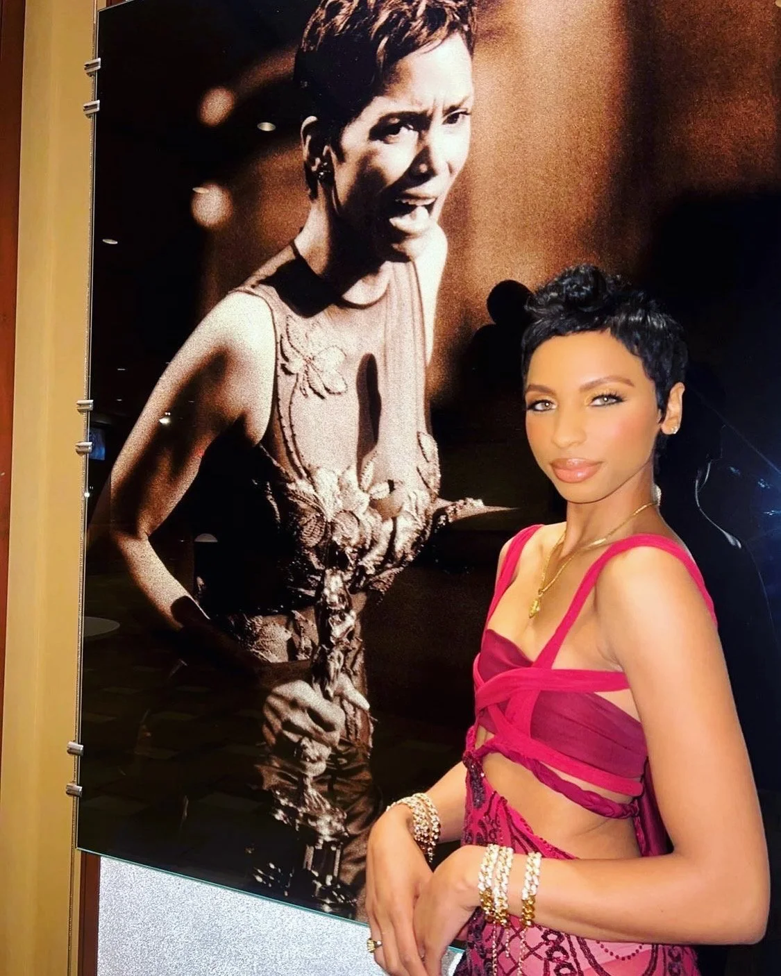 A woman with short black hair and wearing a pink dress with cutout sections and jewelry poses next to a large black and white poster of a woman singing into a microphone.