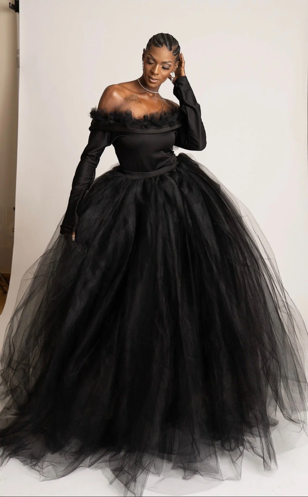 A woman in a long, black, off-the-shoulder gown with a full tulle skirt and fitted bodice, standing against a plain background.