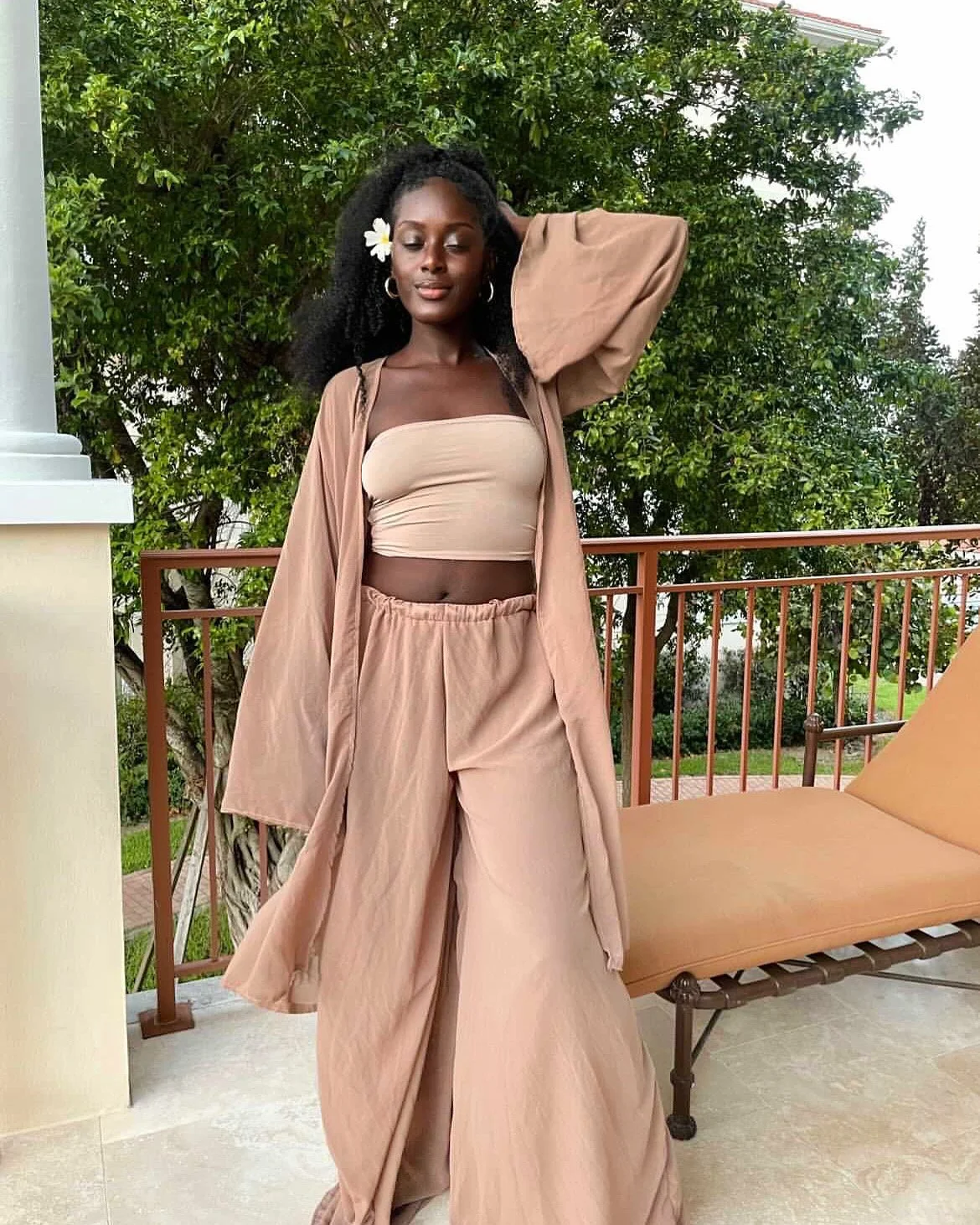 A woman with curly black hair and a flower in her hair, wearing a beige tube top, beige baggy pants with an elastic waistband, and a matching beige long jacket, stands outdoors on a balcony with a greenery background. She has one hand behind her head