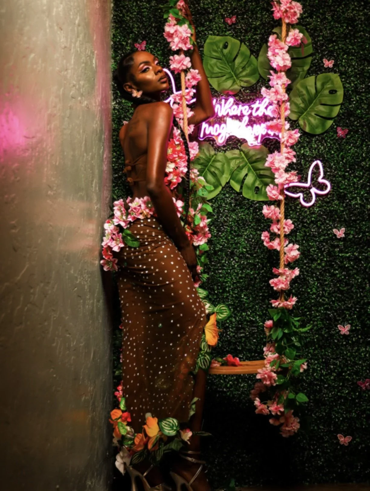 A woman in a sheer brown polka dot skirt and floral top poses against a decorated green wall with pink flowers, large green leaves, and neon pink sign reading 'where the magic happens', surrounded by pink butterflies.