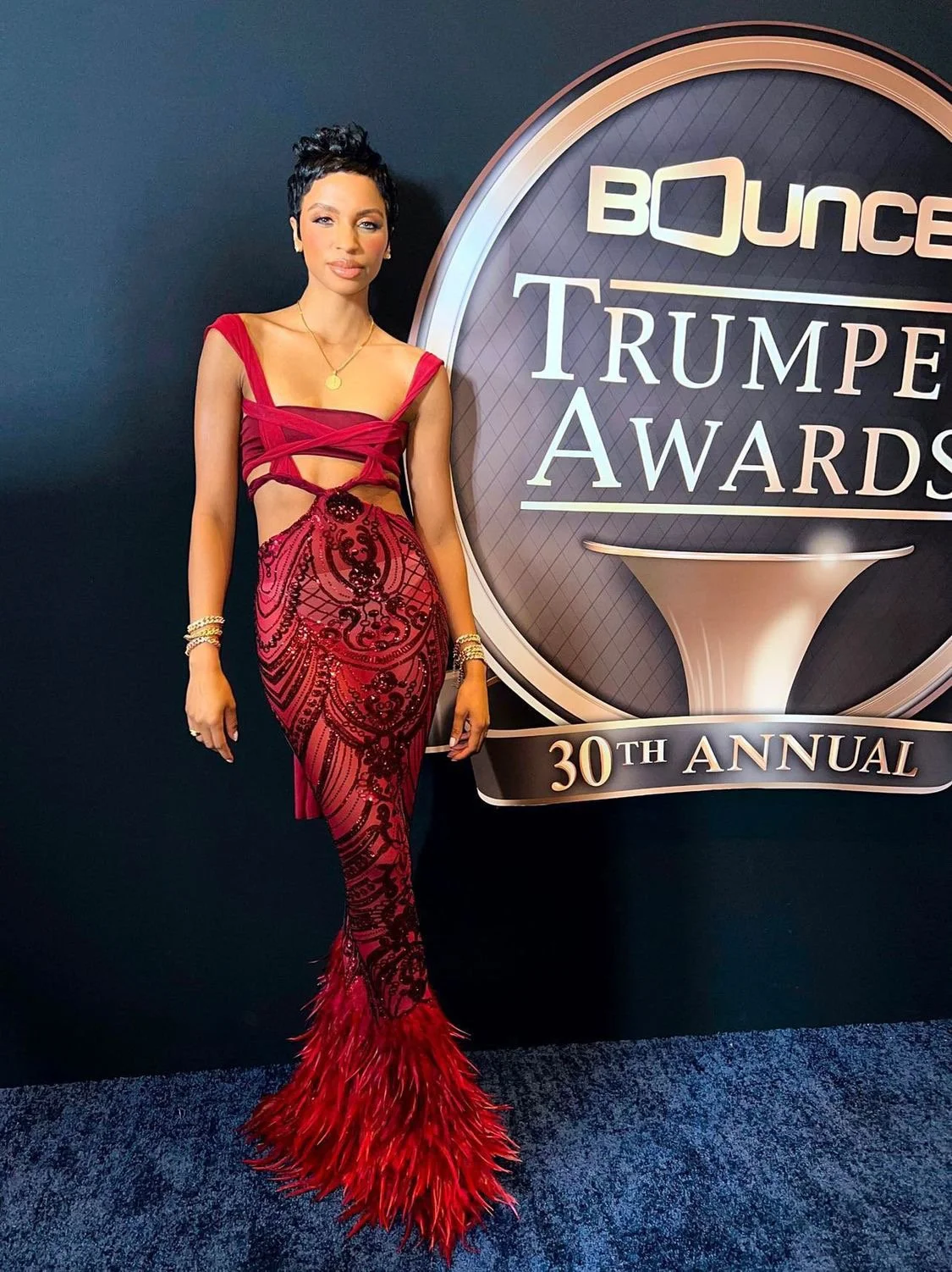 A woman in a red, intricately patterned gown with feathered hemline standing next to a sign that reads 'Bounce Trumpet Awards 30th Annual' at an event.