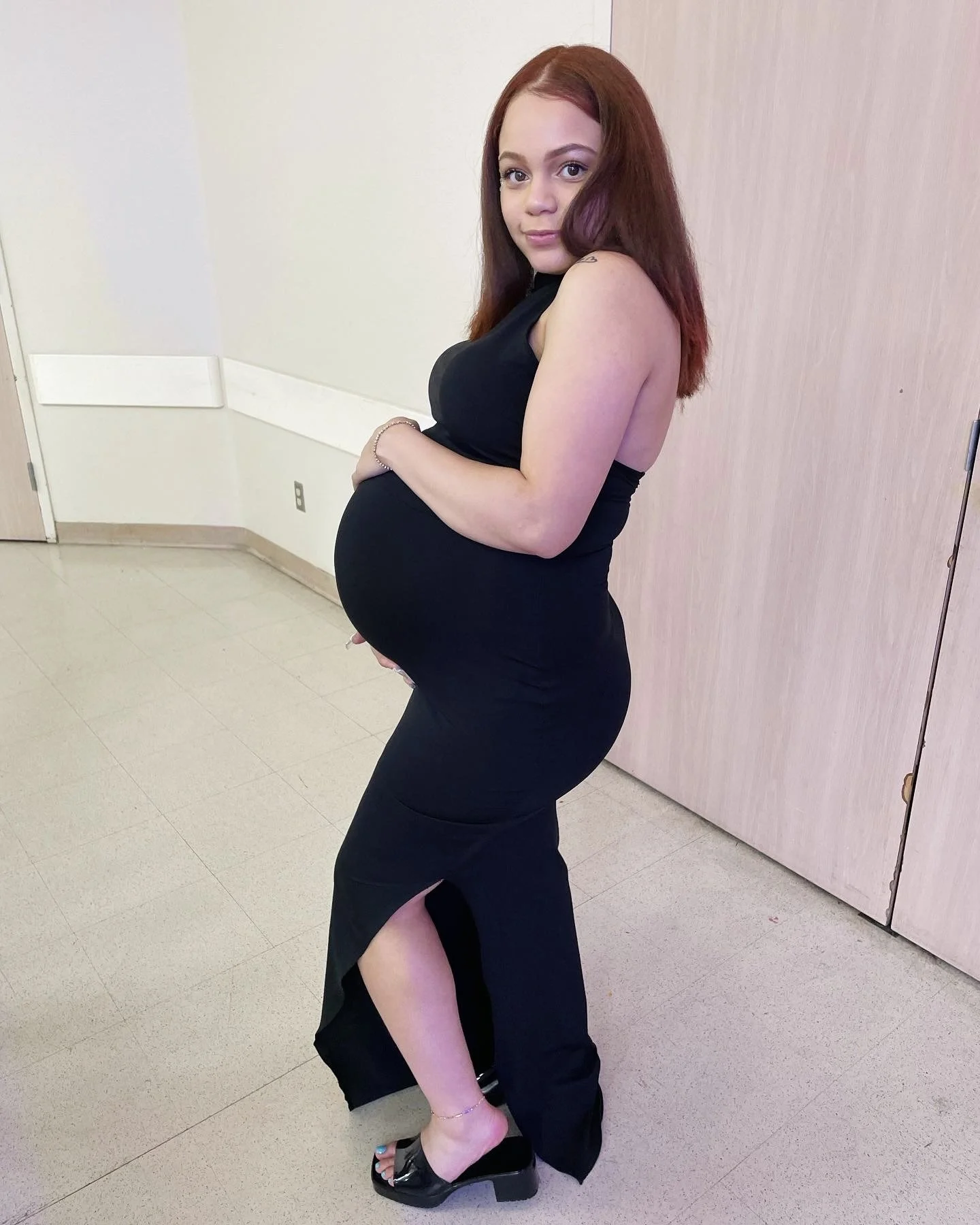A pregnant woman with red hair in a black dress posing in a room with light-colored walls and wooden panels, standing on tiled flooring.