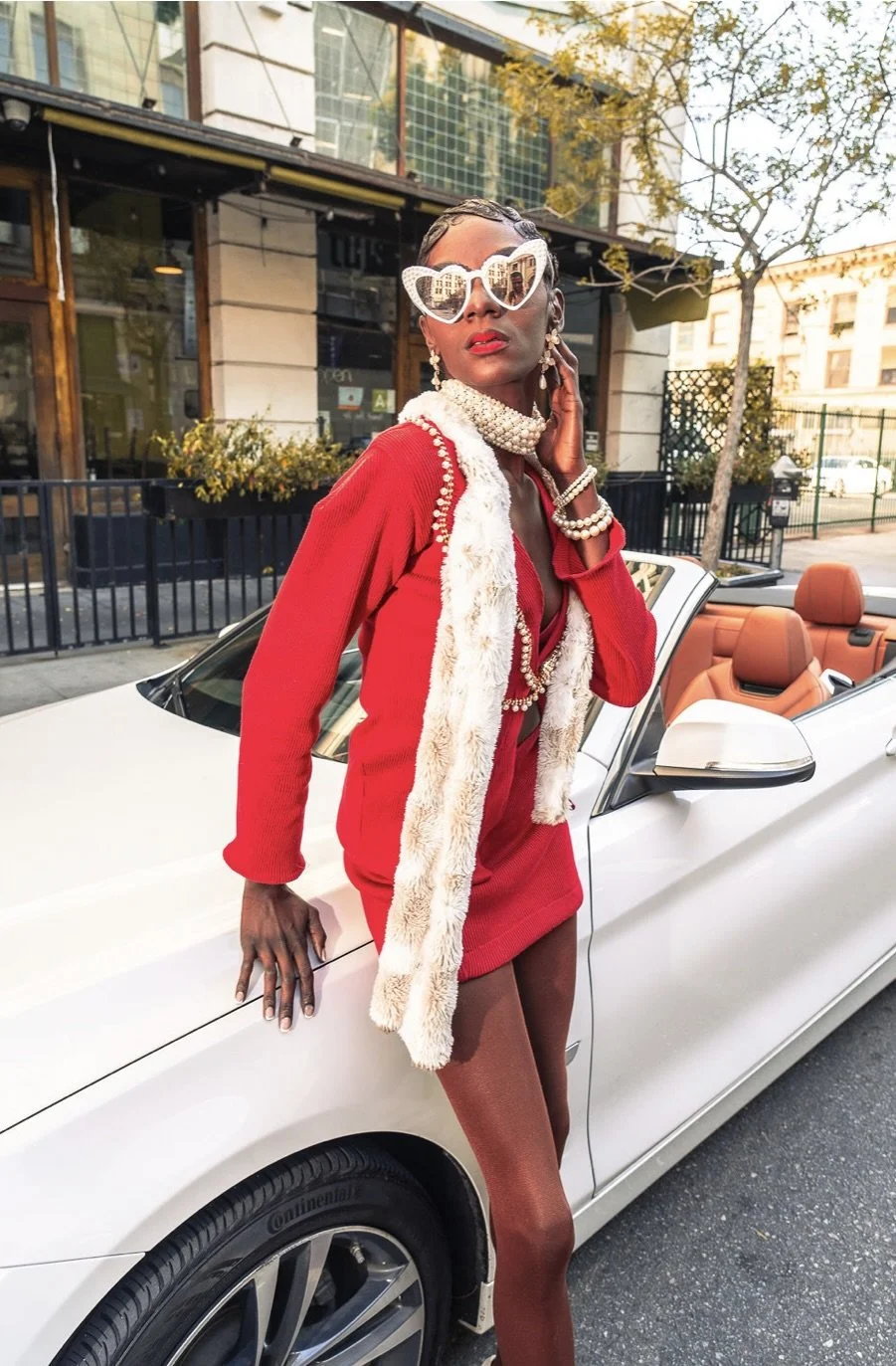 Woman dressed in red with a faux fur stole, wearing large white heart-shaped sunglasses and multiple pearl jewelry, standing next to a white convertible car with tan interior, on a city street.