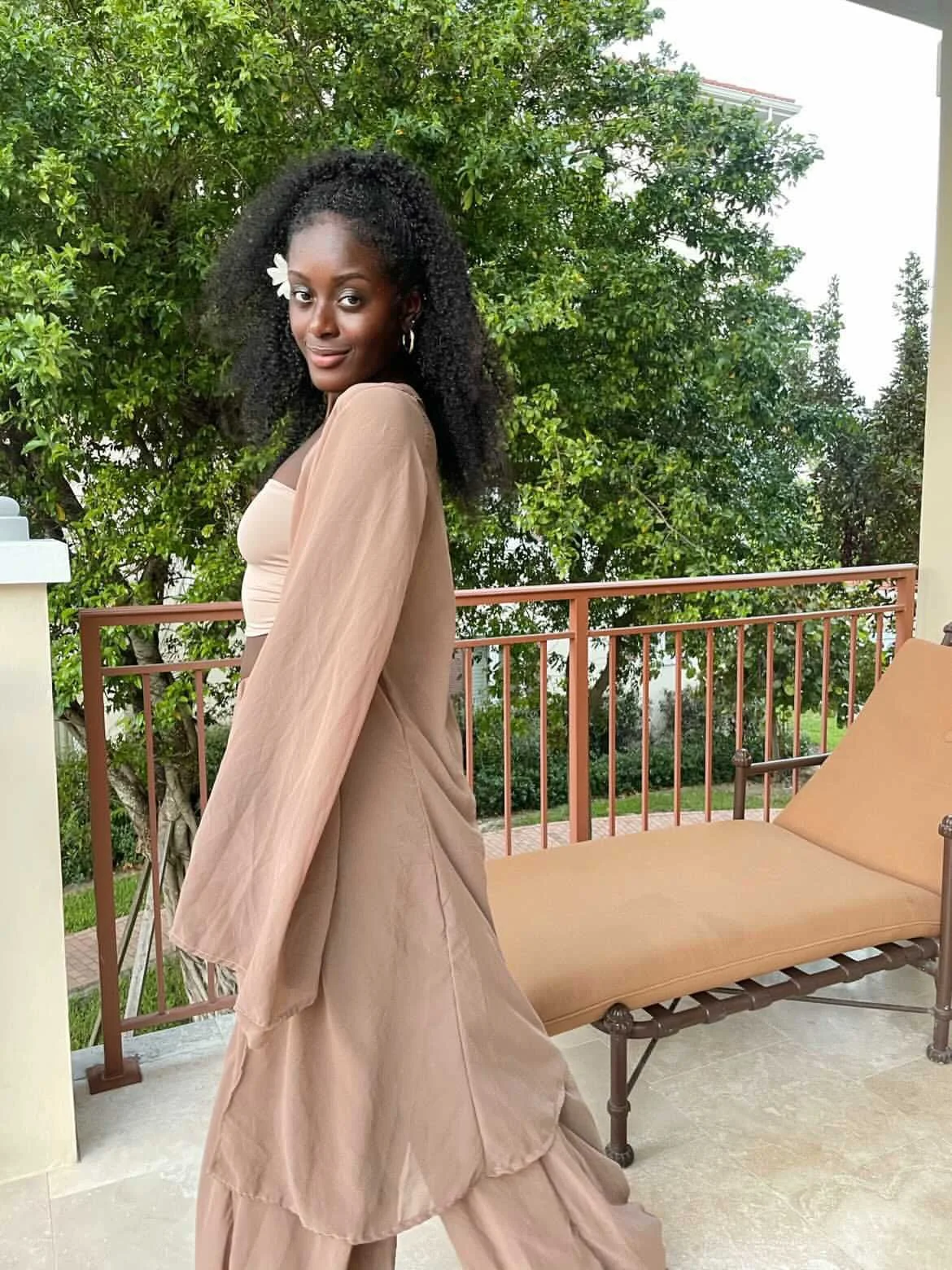 A young woman with dark curly hair wearing a beige outfit and a white flower in her hair, posing on a balcony with green trees in the background.