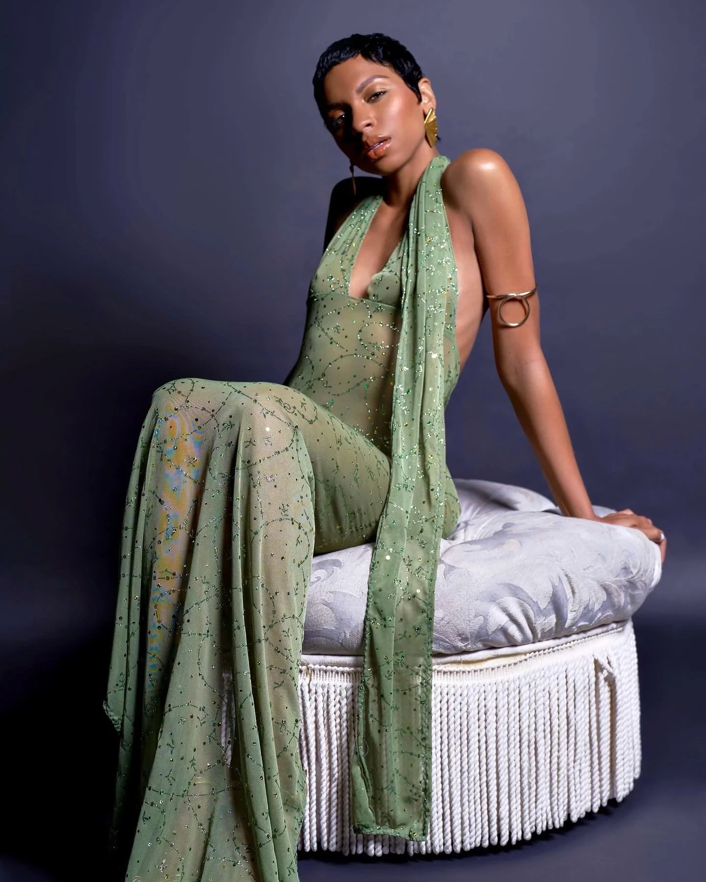A woman with short black hair and gold earrings sits on a white upholstered stool, wearing a sheer, green sequined dress with an open back and a matching scarf draped over her shoulder, against a dark background.