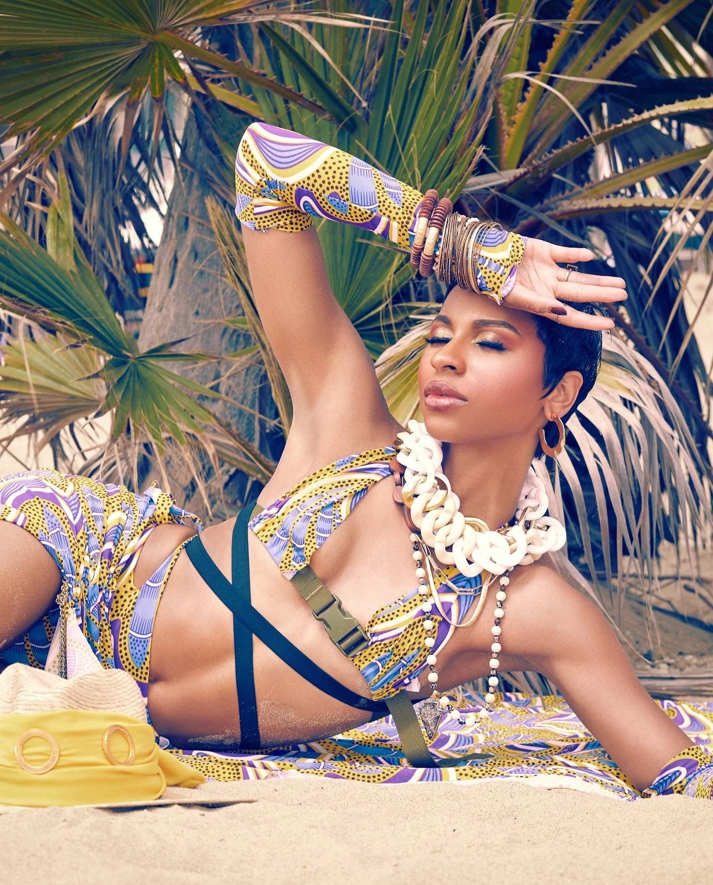 woman wearing colorful patterned swimwear and chunky jewelry reclining on a sandy beach with palm trees in the background