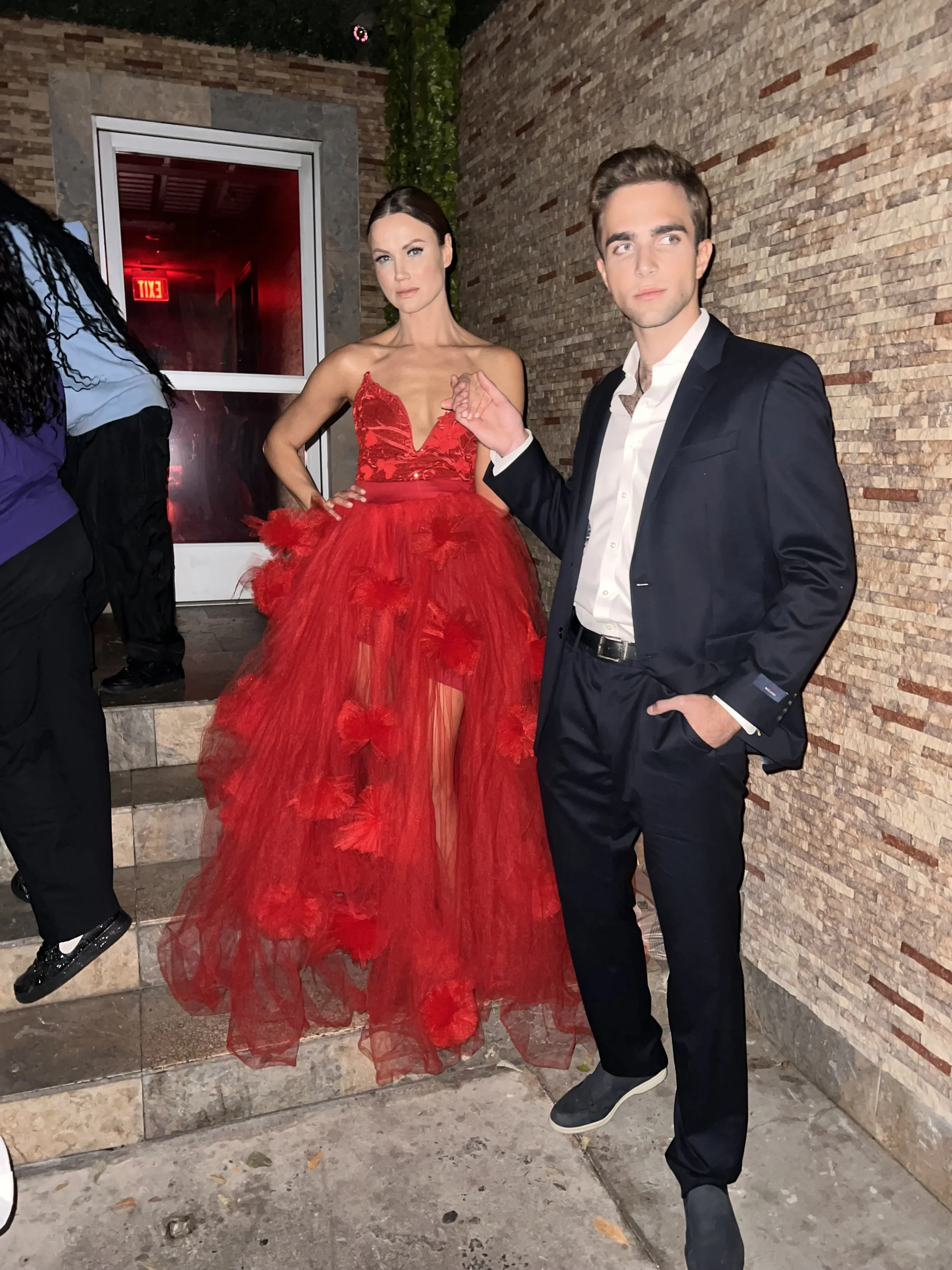 A woman in a red evening gown standing next to a man in a black suit at an indoor event.