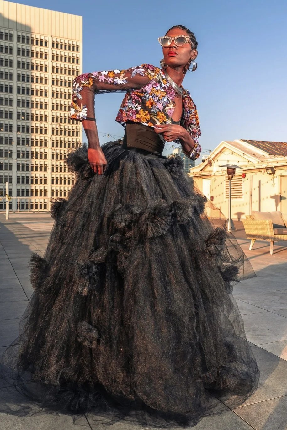 A woman wearing a floral top, large sunglasses, and a black tulle skirt poses outdoors with city buildings in the background during sunset.