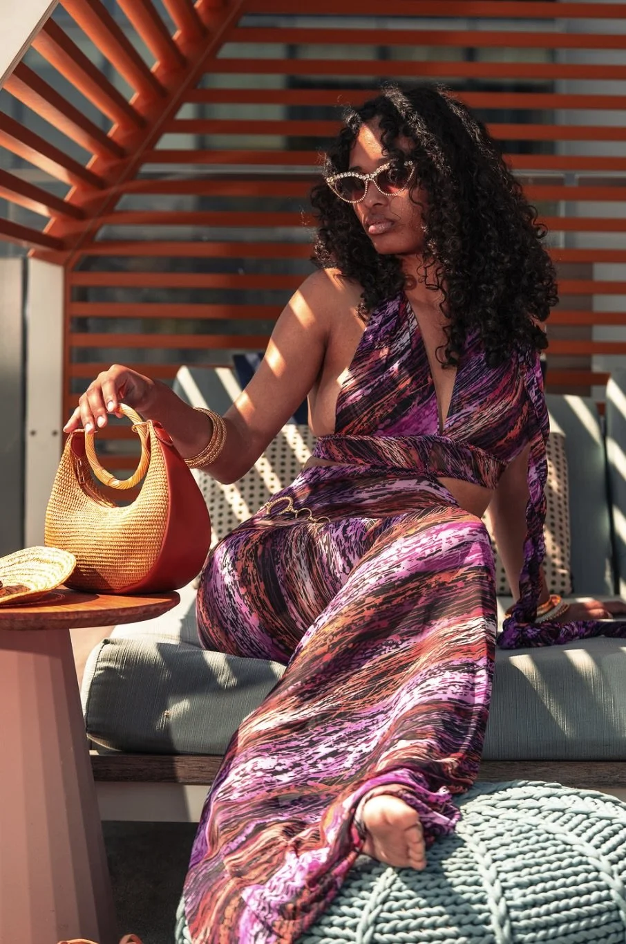 A woman with curly hair wearing a purple patterned halter top and matching pants, sitting outdoors on a cushioned surface, holding a yellow woven handbag, with sunglasses and jewelry, under a wooden slatted pergola.